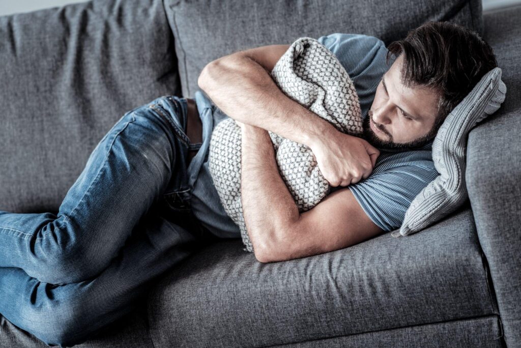 Depressed unhappy sad man lying on the sofa and hugging a cushion while feeling lonely