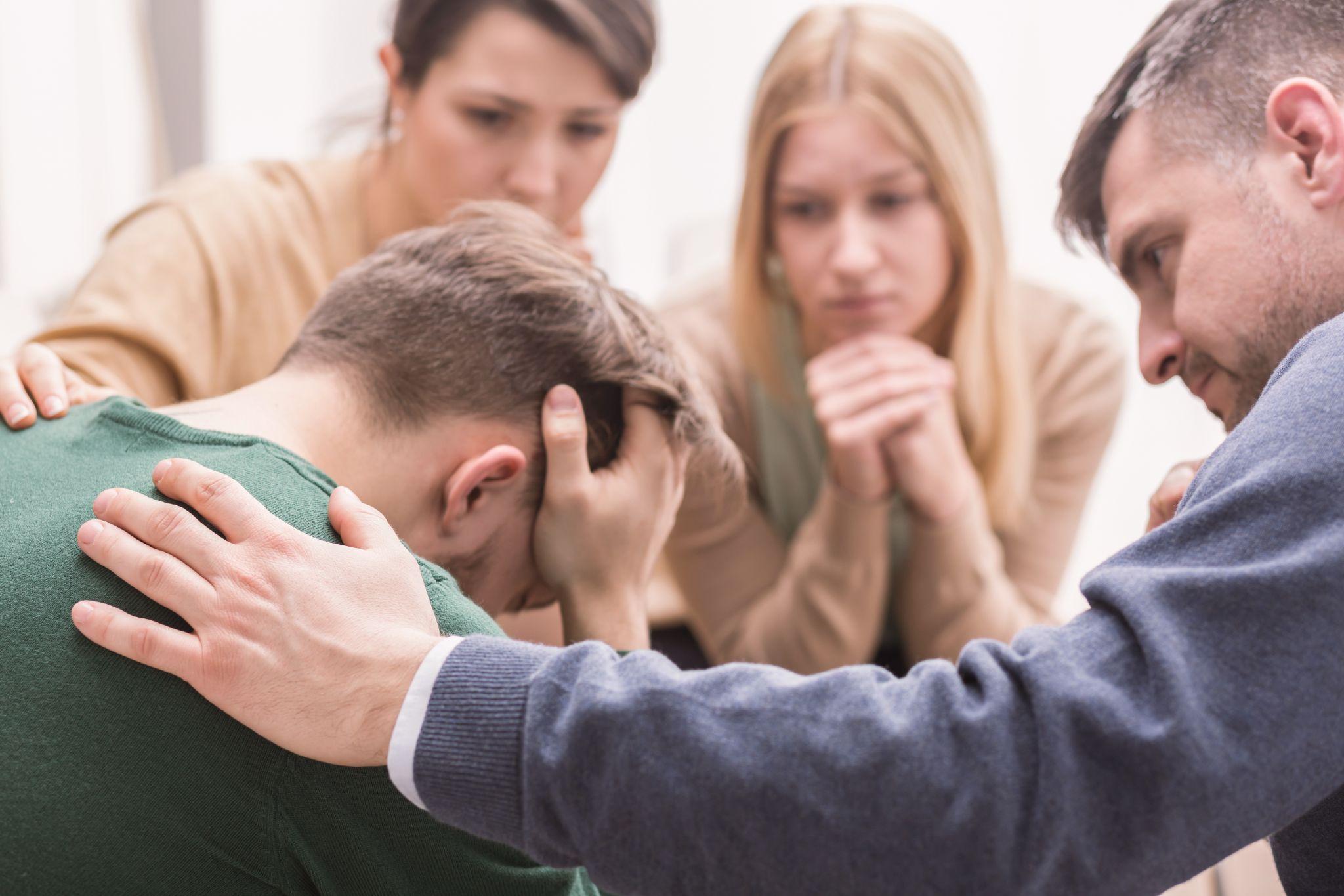Devastated young man holding his head in his hands and friends supporting him during group therapy