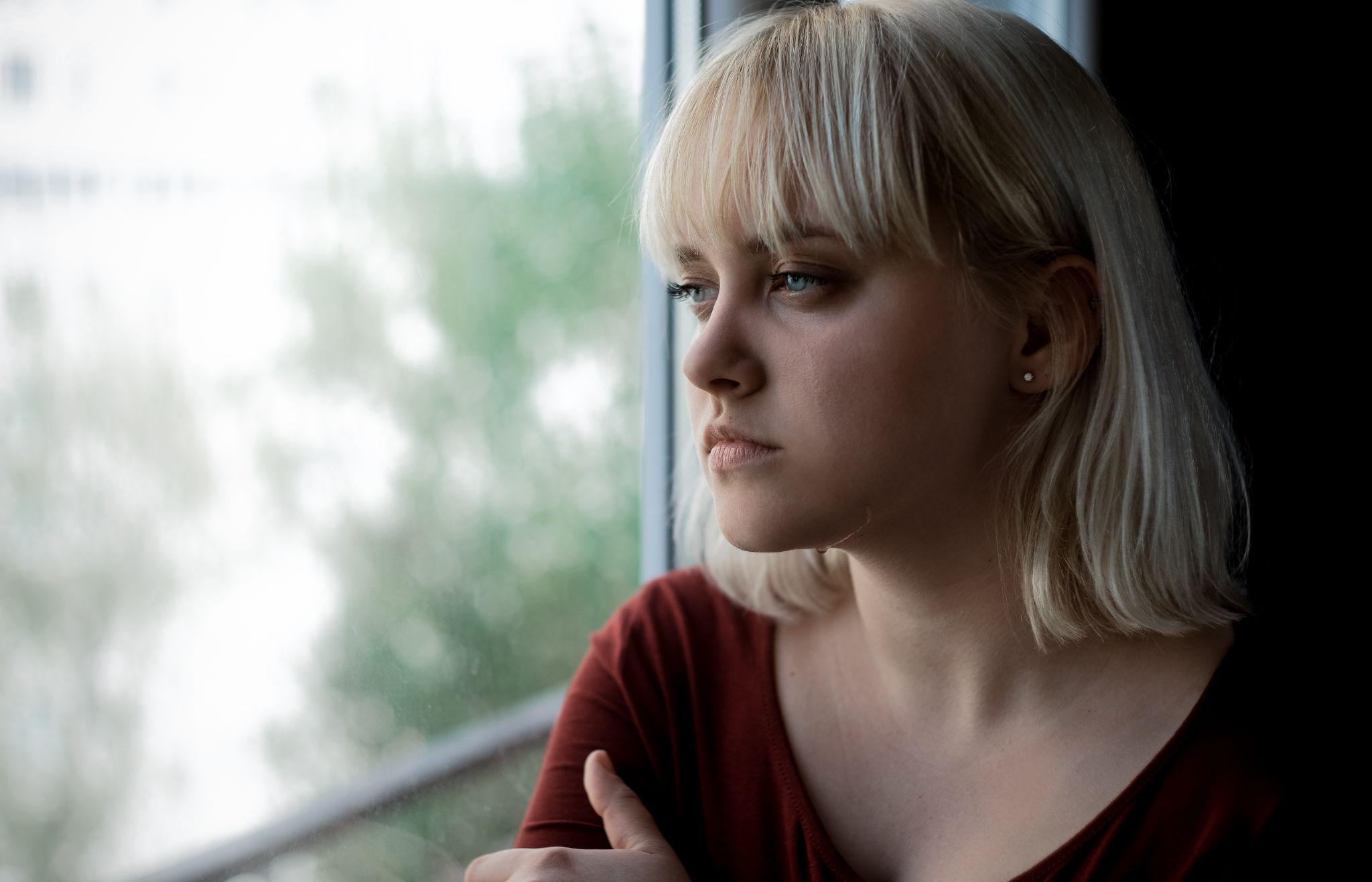 crying depressed young blond woman near window at home.