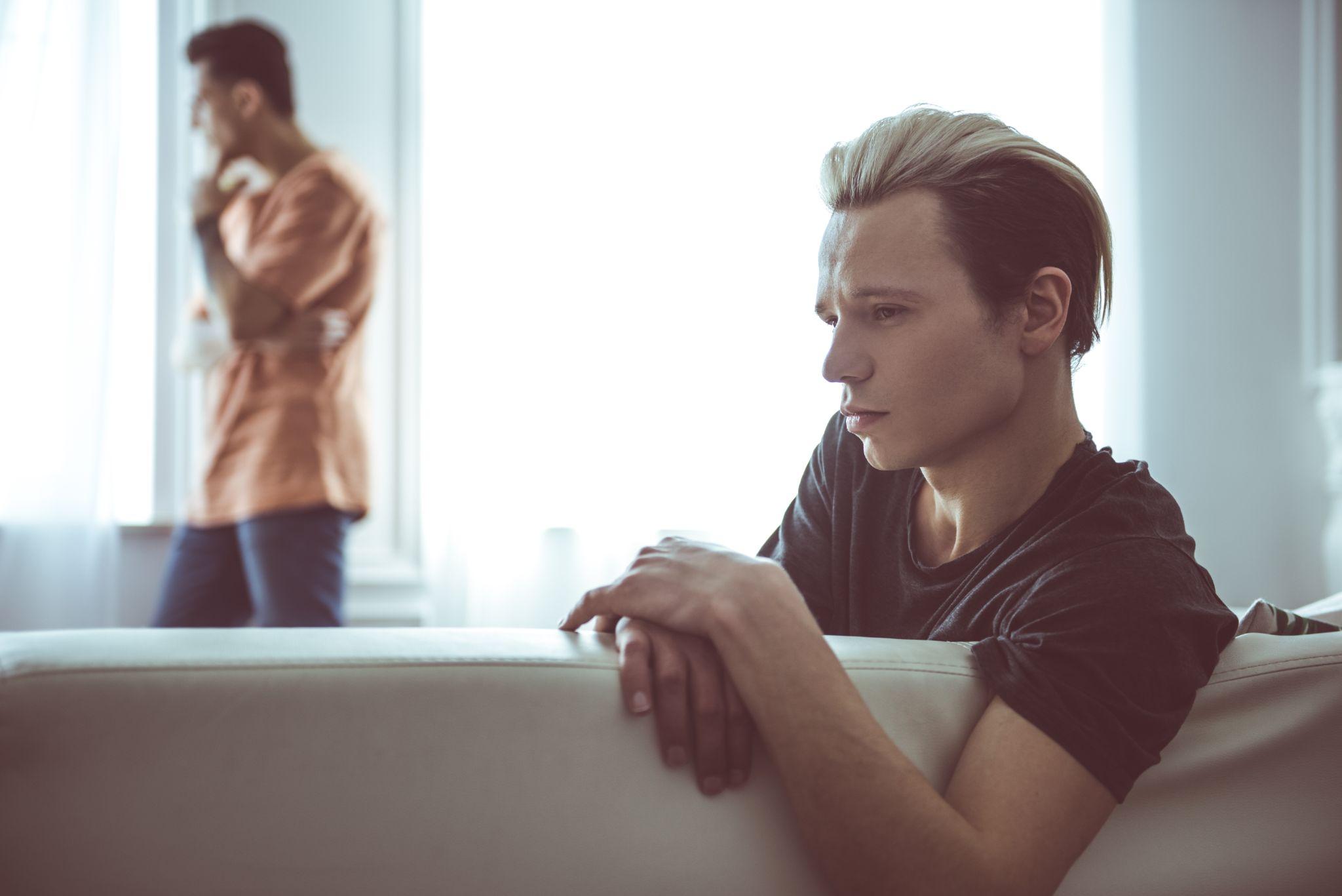 Sad guy with dyed hair sitting on couch while his boyfriend standing near window on blurred