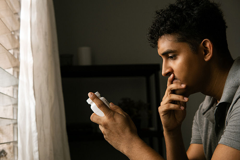 Thoughtful man looking down at pill bottle in dark apartment